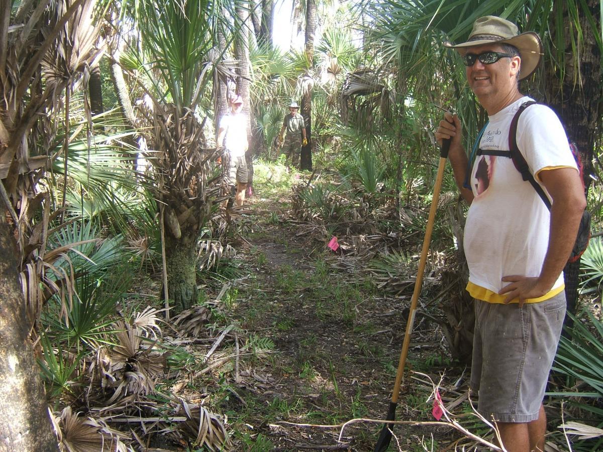 A man stands on a dirt path in a lush green area, surrounded by palm trees and other vegetation, holding a handheld tool. He is wearing a wide-brimmed hat and sunglasses, and he smiles at the camera. In the background, other individuals can be seen working in the same environment, with pink flags marking areas of interest. Carlton Preserve mountain bike trail.