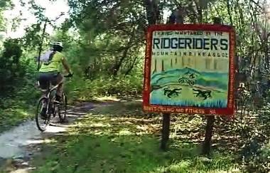 A mountain biker rides along a path in a wooded area, passing a colorful sign that reads "Ridge Riders Mountain Bike Area." The sign features graphics of a cyclist and various animals, indicating a recreational space for mountain biking and fitness activities. Loyce E. Harpe Park mountain bike trail.
