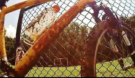 A close-up view of a yellow mountain bike leaning against a black chain-link fence, with a water bottle attached to the frame. The bike is covered in mud, and a grassy field with goalposts is visible in the background under a sunny sky. Loyce E. Harpe Park mountain bike trail.