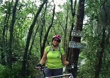 A person wearing a vibrant green shirt and a pink helmet stands next to a trail sign in a lush, wooded area. The sign indicates the names "Baby" and "Beast." The background features tall trees and hanging moss, creating a natural outdoor setting. Loyce E. Harpe Park mountain bike trail.