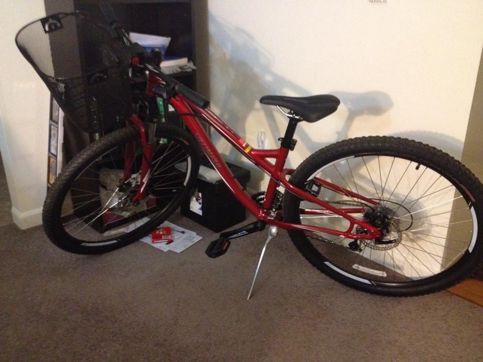 Specialized Myka Sport Disk 29: A red mountain bike with a black front basket is leaning against a bookshelf on a carpeted floor. The bike features thick tires and a simple design, with the seat positioned at a moderate height. Papers and other items are visible on the floor nearby.