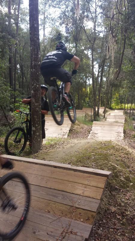 A mountain biker performing a jump over a wooden ramp in a forested area, with another biker and a person watching nearby. The scene features dirt trails and wooden features designed for biking, surrounded by trees. Santos mountain bike trail.