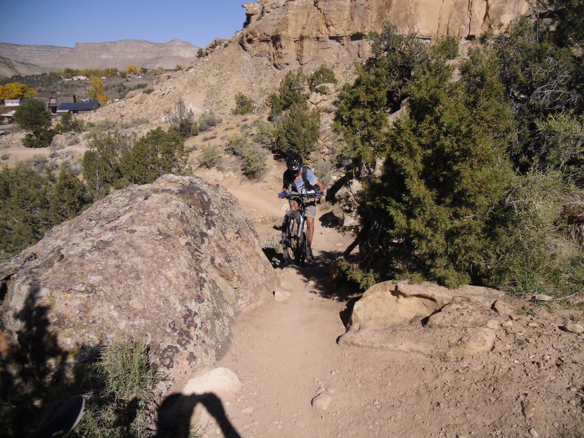 A mountain biker riding along a narrow dirt path, surrounded by rocky terrain and vegetation, with cliffs and distant hills in the background under a clear blue sky. Palisade Rim mountain bike trail.