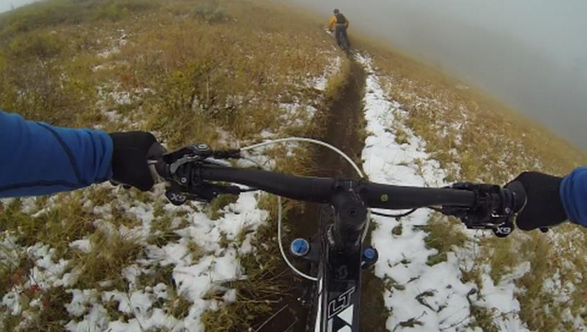 A first-person view of a mountain biking trail, showing the handlebars of a bike and the path ahead, which is partially covered in snow. In the background, a person in an orange jacket is riding on the path through a foggy landscape. Corvair mountain bike trail.