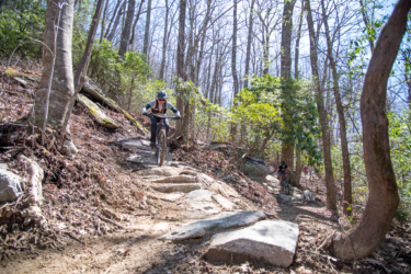 A mountain biker navigates a rocky trail in a wooded area, surrounded by tall trees and greenery. The cyclist is focused on the path ahead, with another rider visible in the background. Sunlight filters through the trees, illuminating the trail. Butter Gap / Trail #123 mountain bike trail.