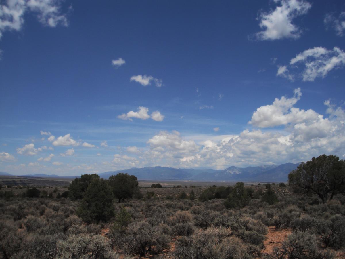 A vast landscape featuring dry terrain with scattered shrubs under a bright blue sky, dotted with white clouds. In the background, mountains are visible, adding depth to the scene. Taos Valley Overlook mountain bike trail.