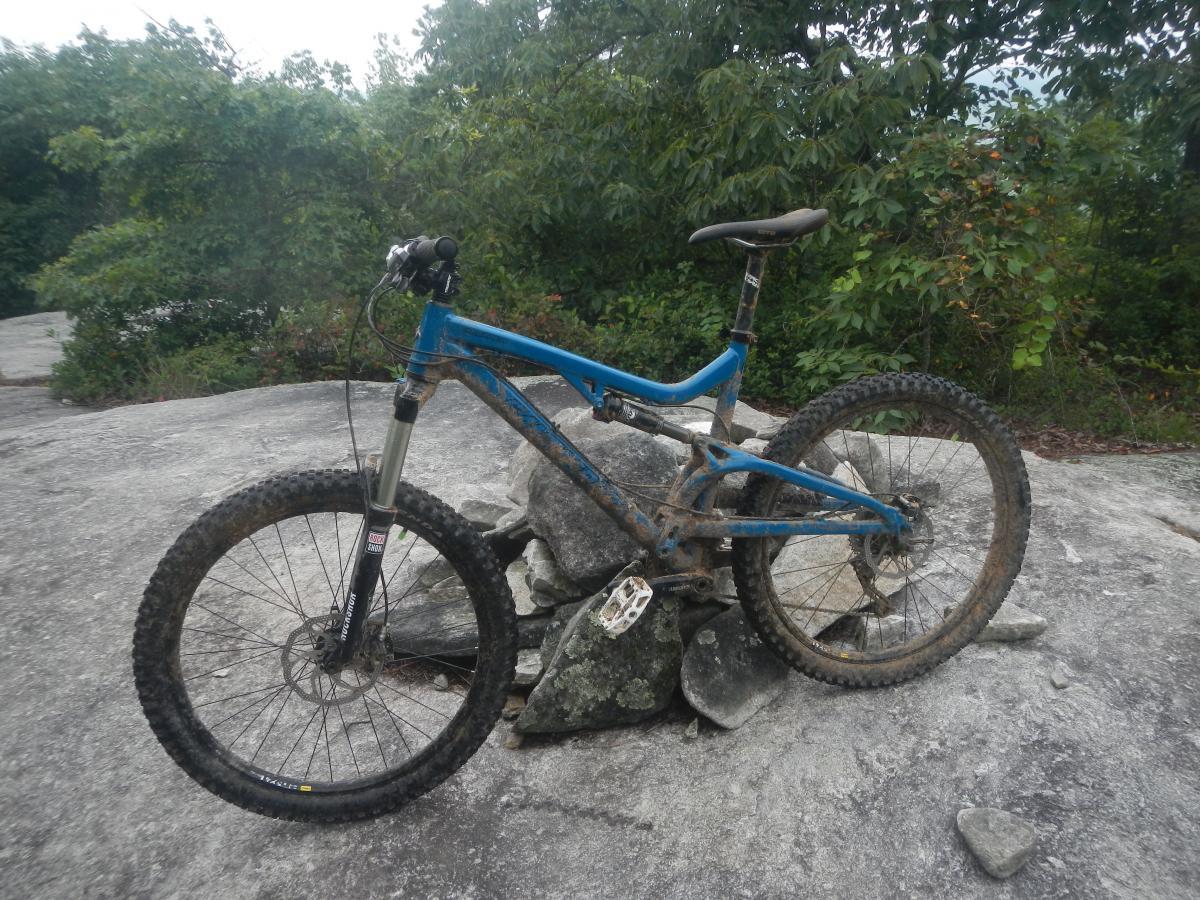 Santa Cruz Heckler: A blue mountain bike rests on a rocky surface, covered in dirt and mud, surrounded by greenery. The bike features a front suspension fork and thick, knobby tires, indicating it has been used for off-road riding. Soft, cloudy skies are visible in the background.