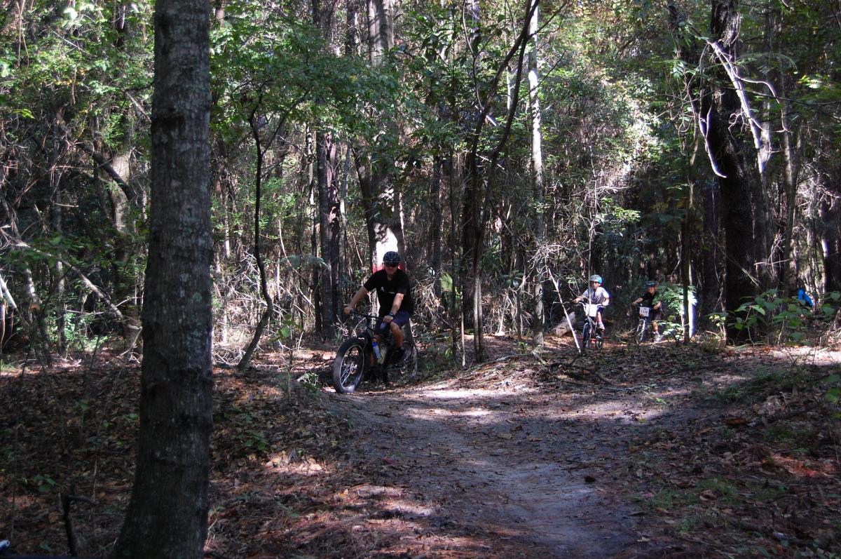 A group of mountain bikers riding on a dirt trail through a dense forest. The scene shows trees and foliage surrounding the path, with sunlight filtering through the branches. One rider is in the foreground, while two others are further along the trail in the background. Tillie Fowler Regional Park mountain bike trail.