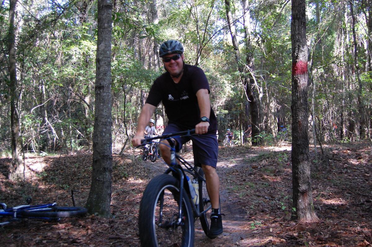 A person riding a mountain bike on a dirt trail surrounded by trees. The scene depicts a sunny day with greenery and a few cyclists visible in the background. The rider is wearing a helmet and sunglasses, smiling as they navigate the trail. Tillie Fowler Regional Park mountain bike trail.