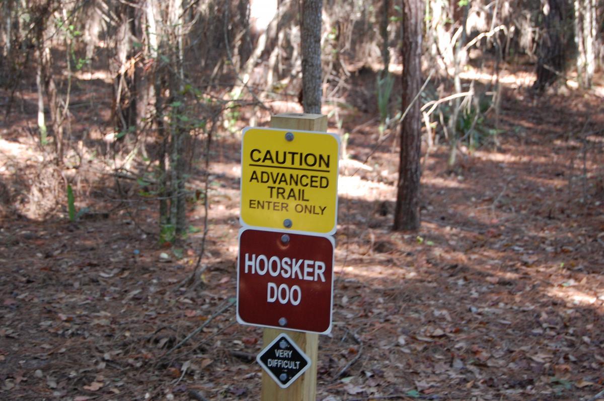 A wooden sign in a wooded area indicating an advanced hiking trail with a warning label. The sign reads "CAUTION ADVANCED TRAIL ENTER ONLY" at the top, followed by "HOOSKER DOO" in bold letters and a smaller sign stating "VERY DIFFICULT." The surrounding environment features trees and scattered fallen leaves. Tillie Fowler Regional Park mountain bike trail.