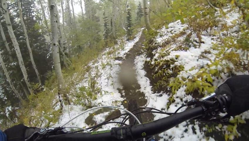 A view from the handlebars of a mountain bike navigating a snowy trail surrounded by tall trees and lush greenery. The ground is partially covered in snow, indicating recent weather changes. TG mountain bike trail.