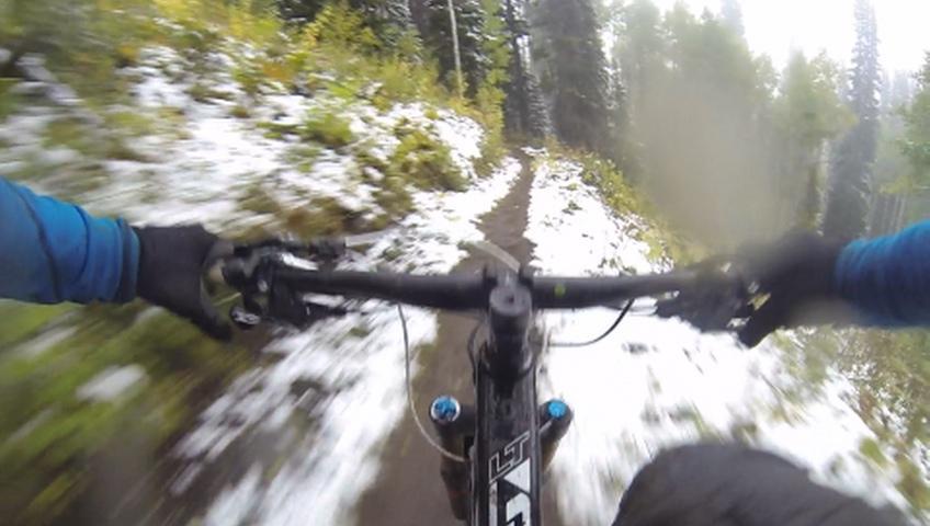 A close-up view of a mountain biker's hands gripping the handlebars as they navigate a snowy and muddy trail in a forested area. The perspective gives a sense of speed and motion, with greenery and a dusting of snow visible along the path. Mid Mountain mountain bike trail.