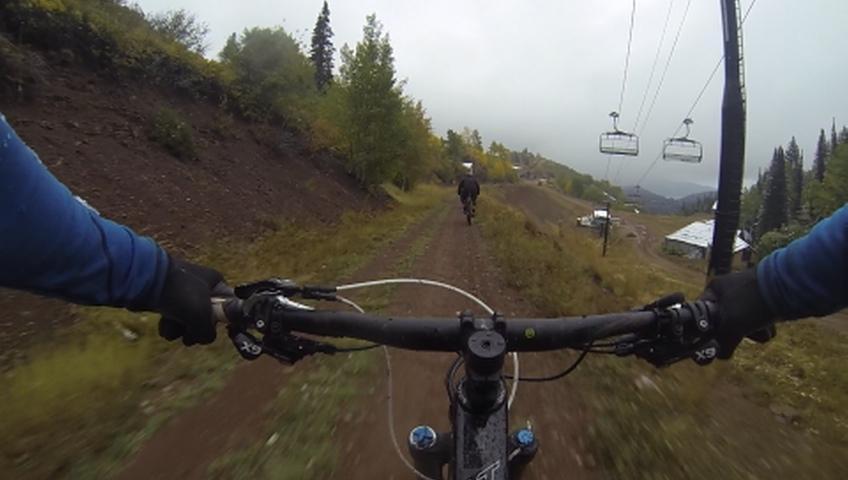 A close-up view from the handlebars of a mountain bike riding down a dirt trail with a rider ahead. The landscape features trees with autumn foliage and a ski lift visible in the background under a cloudy sky. Mid Mountain mountain bike trail.