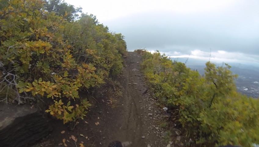 A narrow dirt trail winds through vibrant autumn foliage, with lush green and orange leaves on either side. The trail leads towards a misty view in the distance, partially obscured by clouds. The ground is rocky and uneven, suggesting a rugged hiking or biking path. Mid Mountain mountain bike trail.