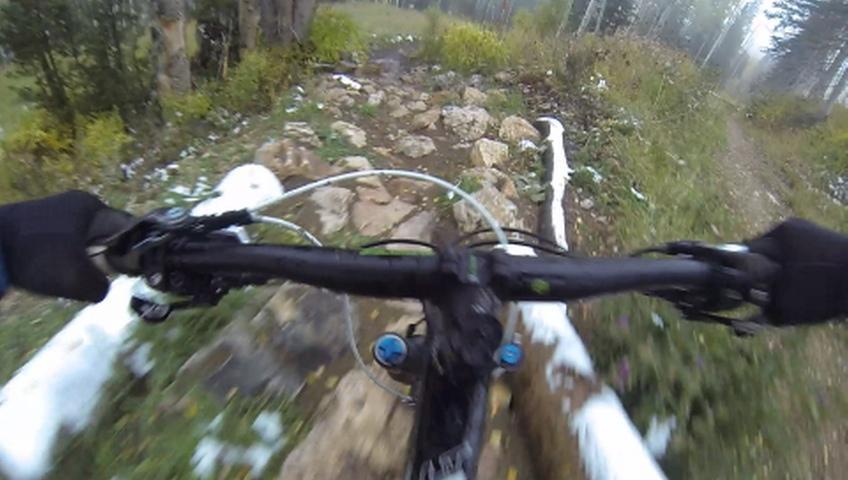 Close-up view of a mountain bike handlebar navigating a rocky trail, with patches of snow and greenery visible along the sides. The perspective suggests an active ride through a forested area on a misty day. Mid Mountain mountain bike trail.