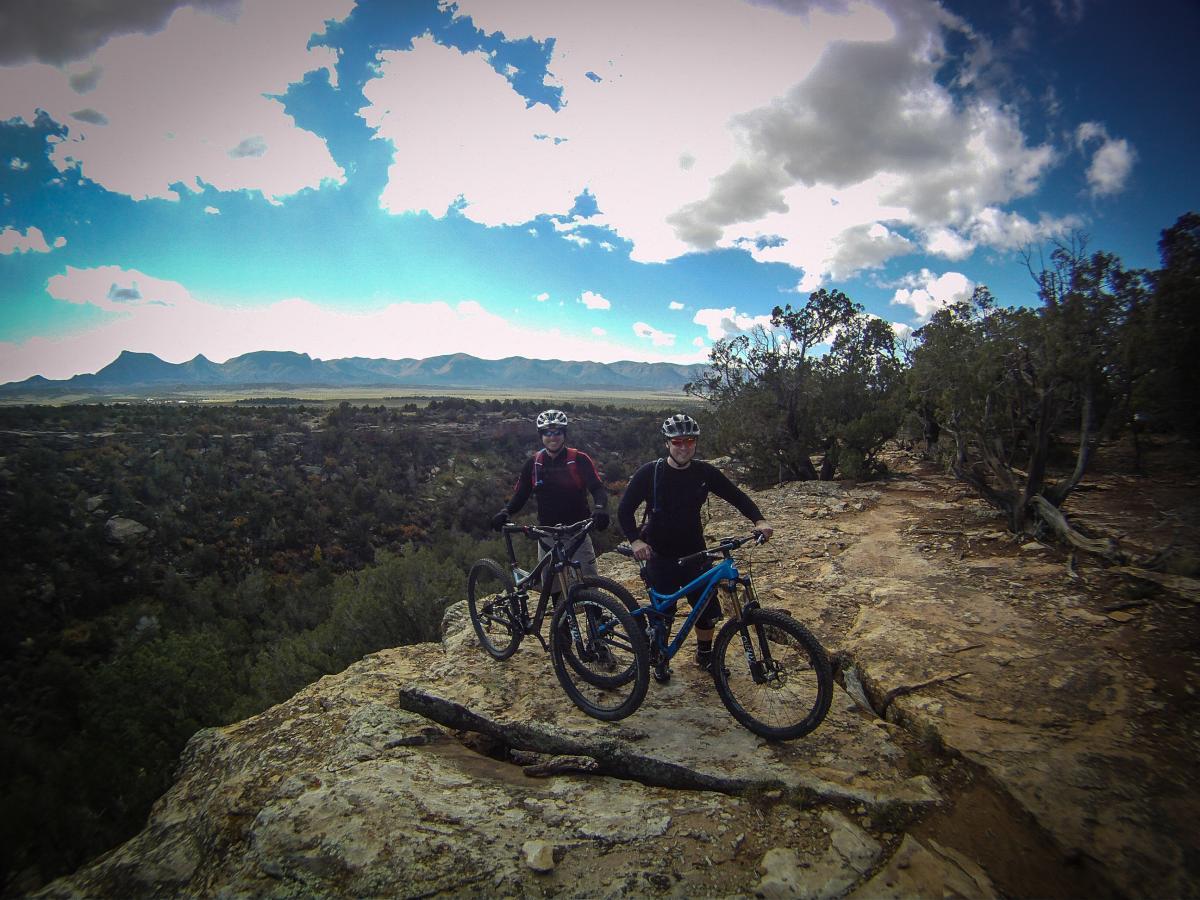 Two mountain bikers standing on a rocky outcrop, surrounded by a scenic landscape with mountains in the background. The sky is partly cloudy, showcasing a blue hue, while lush greenery and rugged terrain can be seen below. One rider is on a black bike and the other on a blue bike, both wearing helmets and cycling gear. Phil's World mountain bike trail.