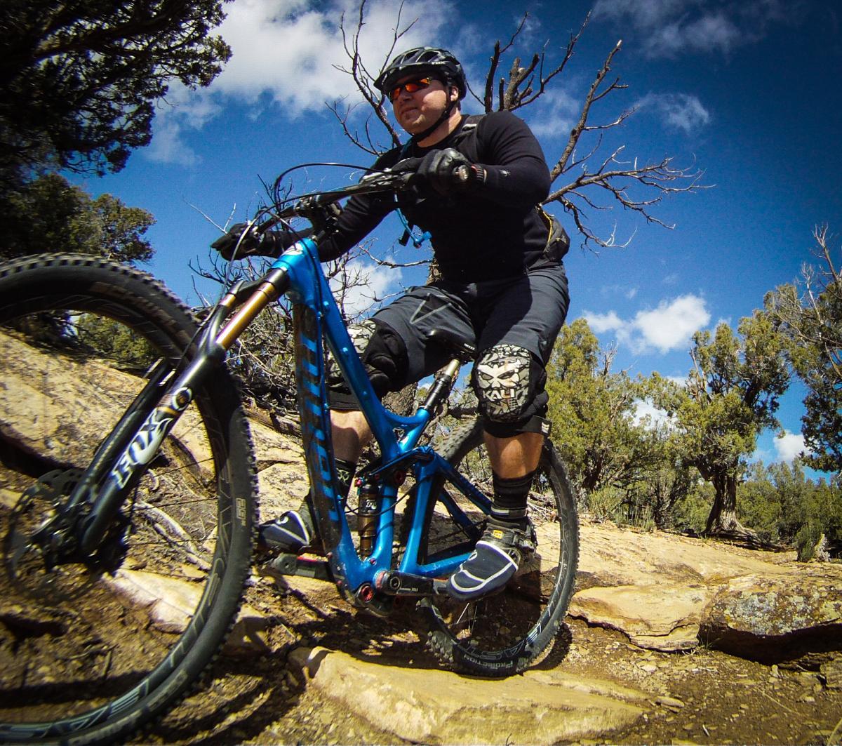 A cyclist riding a blue mountain bike on rocky terrain under a partly cloudy sky. The rider is wearing a black helmet, sunglasses, and protective gear. Surrounding the trail are trees and sparse vegetation, suggesting a rugged outdoor setting. Phil's World mountain bike trail.