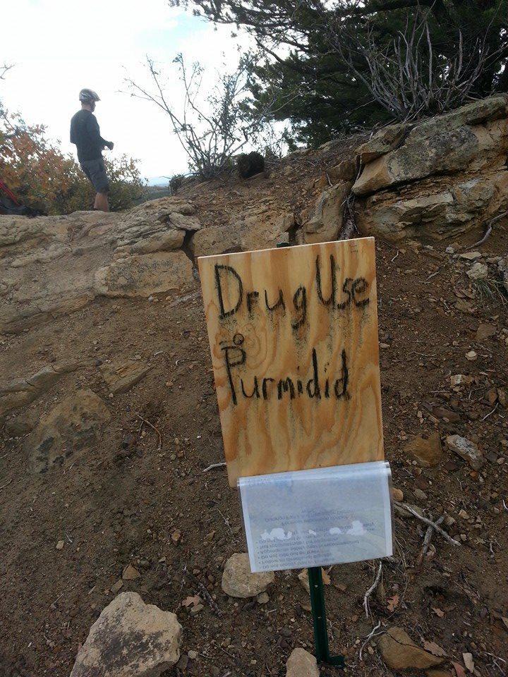 A wooden sign standing on a rocky trail, with the words "Drug Use is Purmidid" written on it. In front of the sign, there is a sheet of paper in a plastic holder, and a person is visible in the background, standing on the path. Surrounding the area are sparse bushes and trees under a cloudy sky. Horse Gulch mountain bike trail.