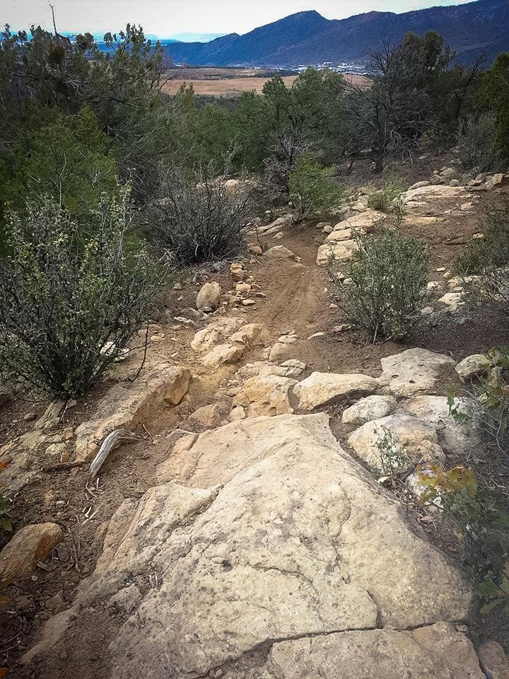 A rocky trail winding through lush vegetation, with large boulders and scattered shrubs, leading down towards a valley and distant mountains under a cloudy sky. Horse Gulch mountain bike trail.
