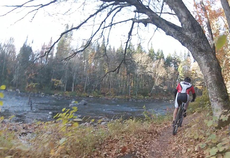 A cyclist rides along a dirt path beside a river, surrounded by trees with autumn foliage. The scene captures the beauty of nature in fall, with fallen leaves lining the trail and a peaceful waterway in the background. Grande Ourse mountain bike trail.