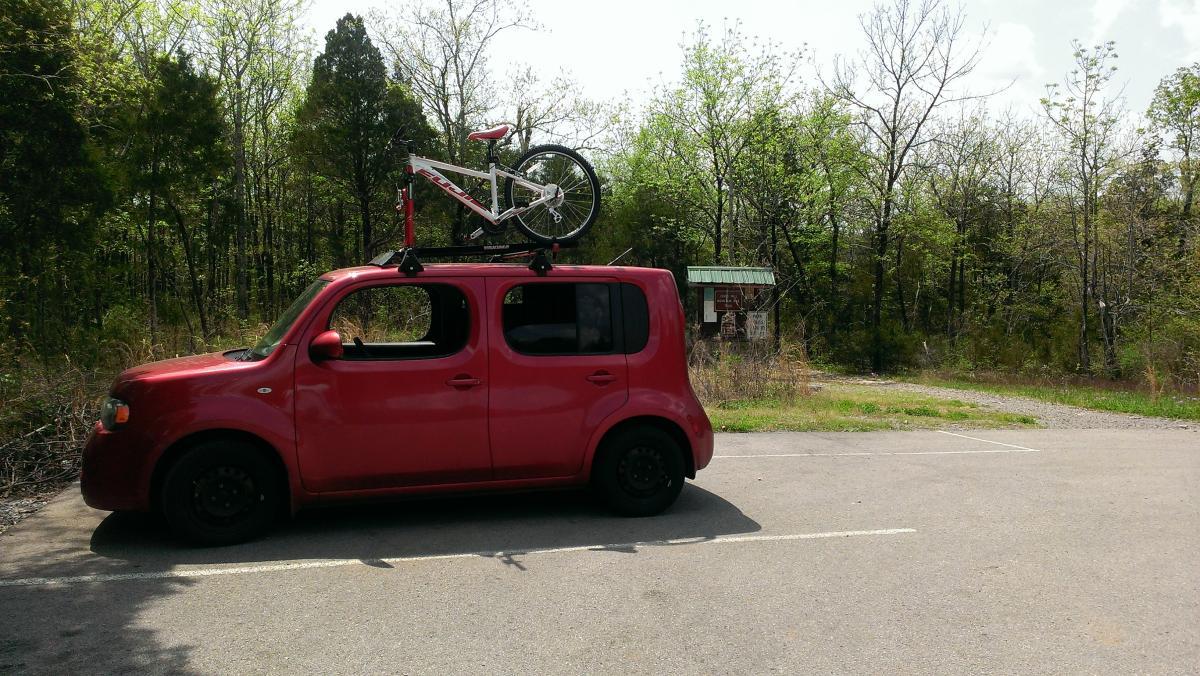 Fuji Nevada 3.0: A red vehicle parked in a lot, with a white bicycle mounted on its roof rack. In the background, there are trees and a small building with a green roof, indicating a natural or recreational area. The scene is set on a sunny day with a clear sky.