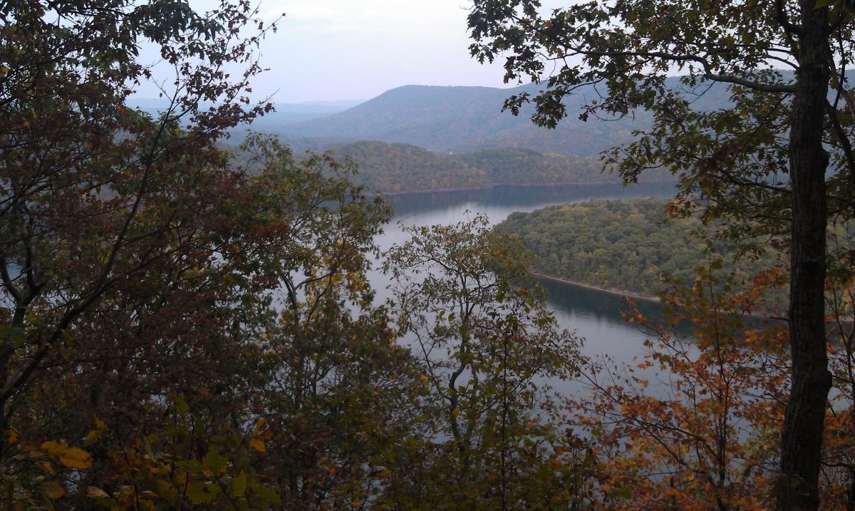 A serene view of a lake surrounded by lush, colorful trees in autumn, framed by leaves in the foreground and mountains in the background under a cloudy sky. Allegrippis Trails mountain bike trail.