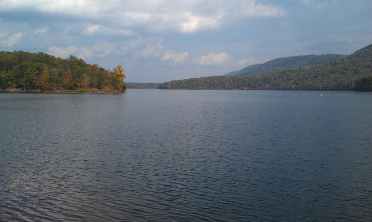 A serene lake view surrounded by autumn-colored trees, with gentle ripples on the water's surface and distant hills under a partly cloudy sky. Allegrippis Trails mountain bike trail.