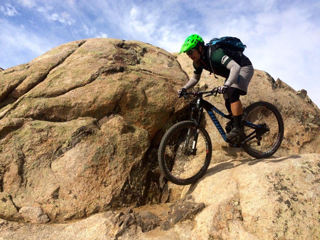 A mountain biker navigating over a rocky terrain, wearing a bright green helmet and gray athletic gear, with a backpack on. The scene captures the rugged landscape with large boulders under a blue sky, showcasing the adventurous spirit of mountain biking. Frontside mountain bike trail.