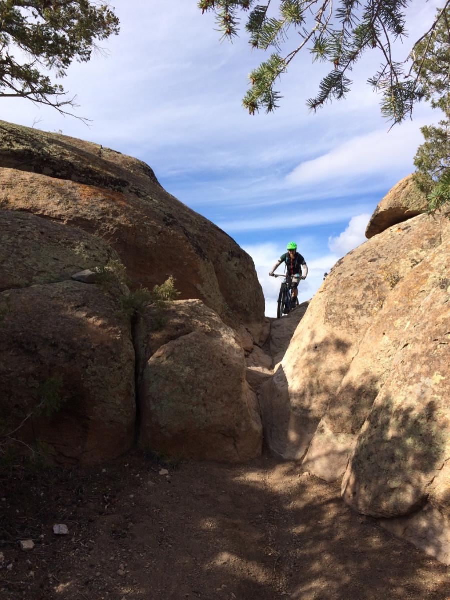 A mountain biker navigating a narrow path between large rocks, surrounded by trees under a partly cloudy sky. Hartman Rocks mountain bike trail.