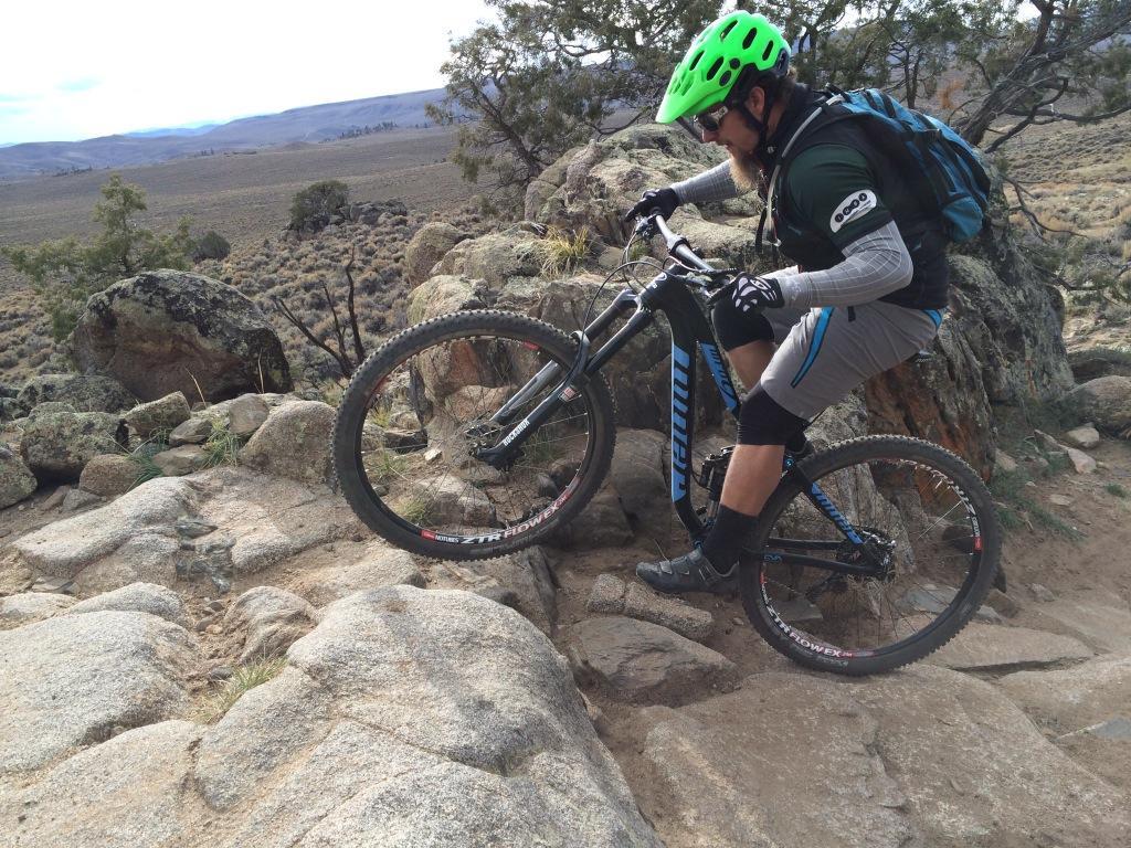 A mountain biker wearing a green helmet and sunglasses navigates rocky terrain, lifting the front wheel of a black and blue mountain bike over a boulder. Surrounding him are sparse bushes and a vast, open landscape in the background. Hartman Rocks mountain bike trail.