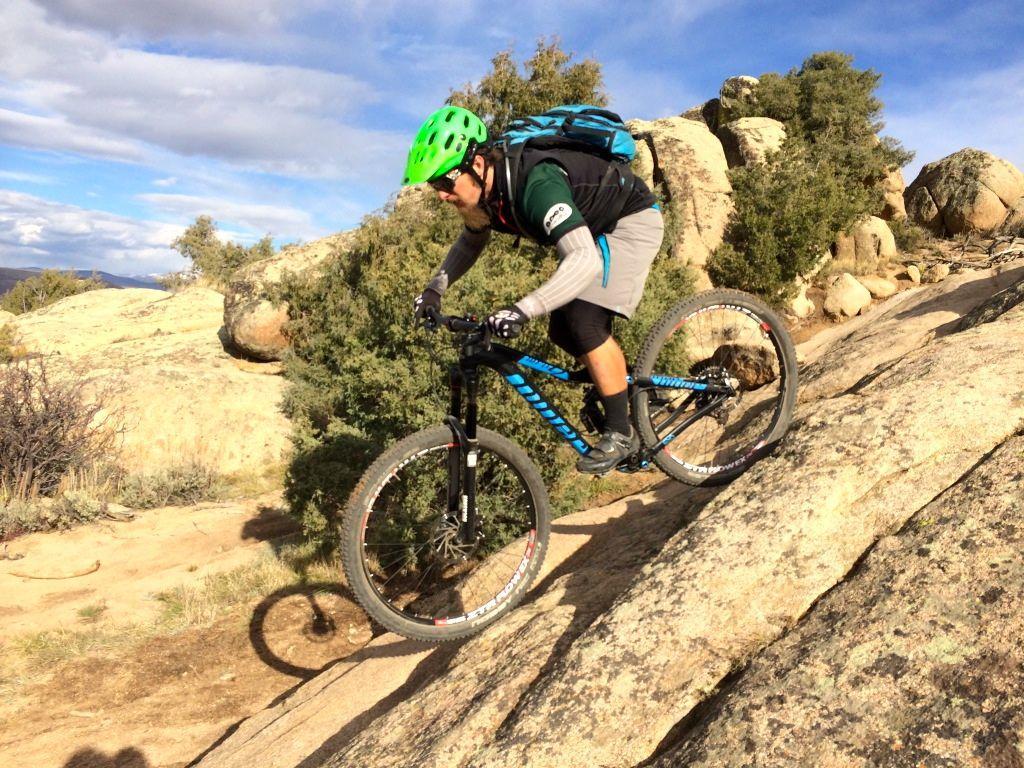 A mountain biker in a green helmet navigates a rocky downhill trail, focused on maintaining balance. The landscape features boulders and sparse vegetation under a partly cloudy sky. Rattlesnake mountain bike trail.