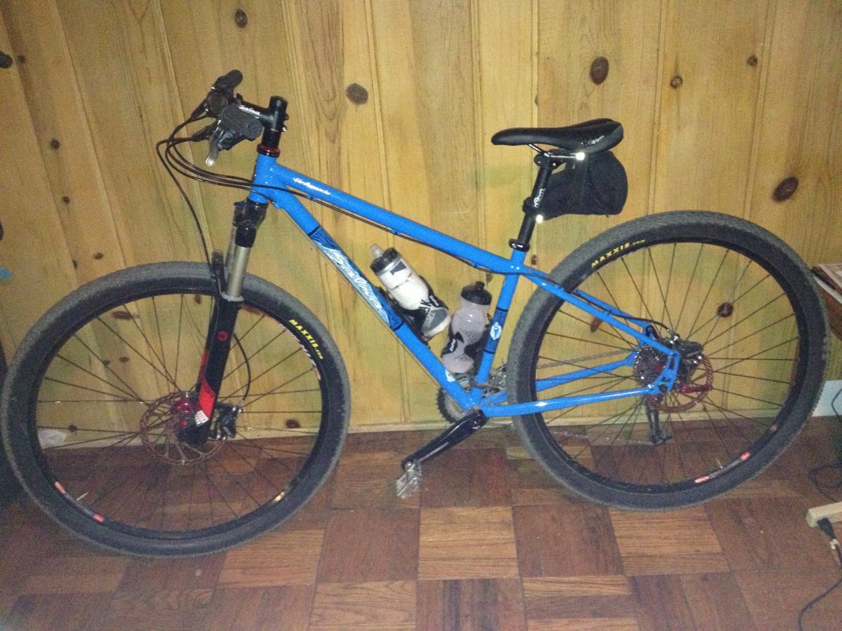 Salsa El Mariachi: A blue mountain bike with a black front fork, featuring thick tires and disc brakes. It has a water bottle mounted on the frame and a small black bag attached under the seat. The bike is positioned against a wooden wall with a parquet floor in the background.