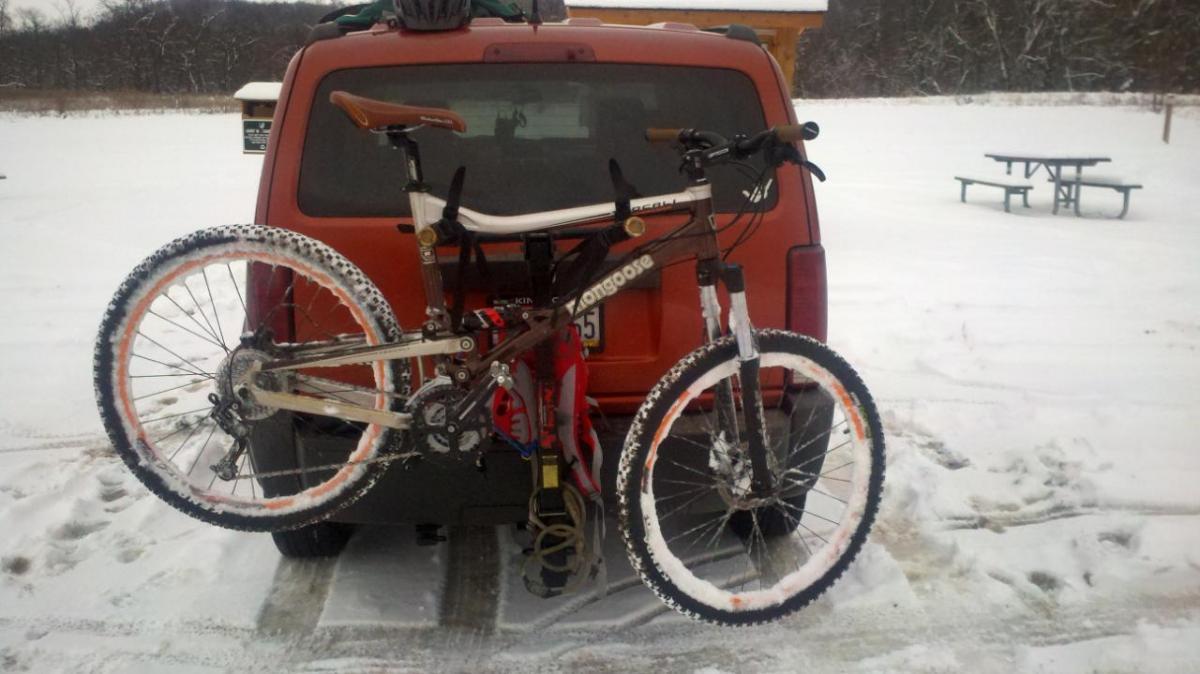 Mongoose Teocali Comp: A mountain bike attached to the back of a vehicle, set in a snowy landscape. The bike features white tires with orange accents, and a dark brown frame. The rear of the vehicle is orange, with a snow-covered ground and a picnic area visible in the background.