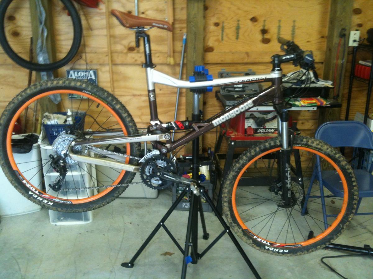 Mongoose Teocali Comp: A mountain bike with a brown and silver frame is mounted on a repair stand in a garage. The bike features orange rims and a leather saddle. The background includes tools and storage items commonly found in a workshop setting.
