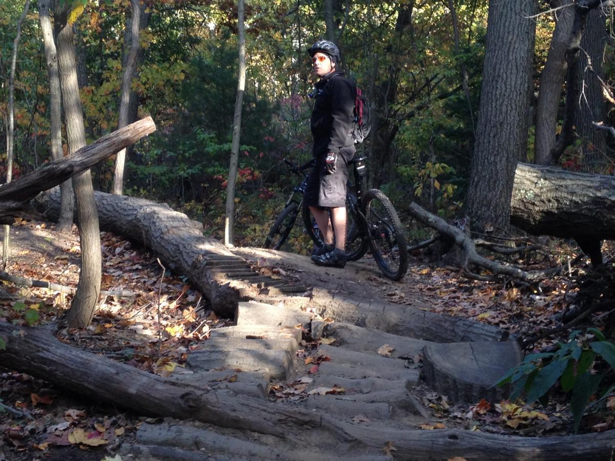 A mountain biker stands next to his bike on a forest trail surrounded by trees and fallen leaves. The trail features a series of wooden steps and a log bridge. The sunlight filters through the foliage, creating a serene outdoor setting. Meadowlark Park mountain bike trail.