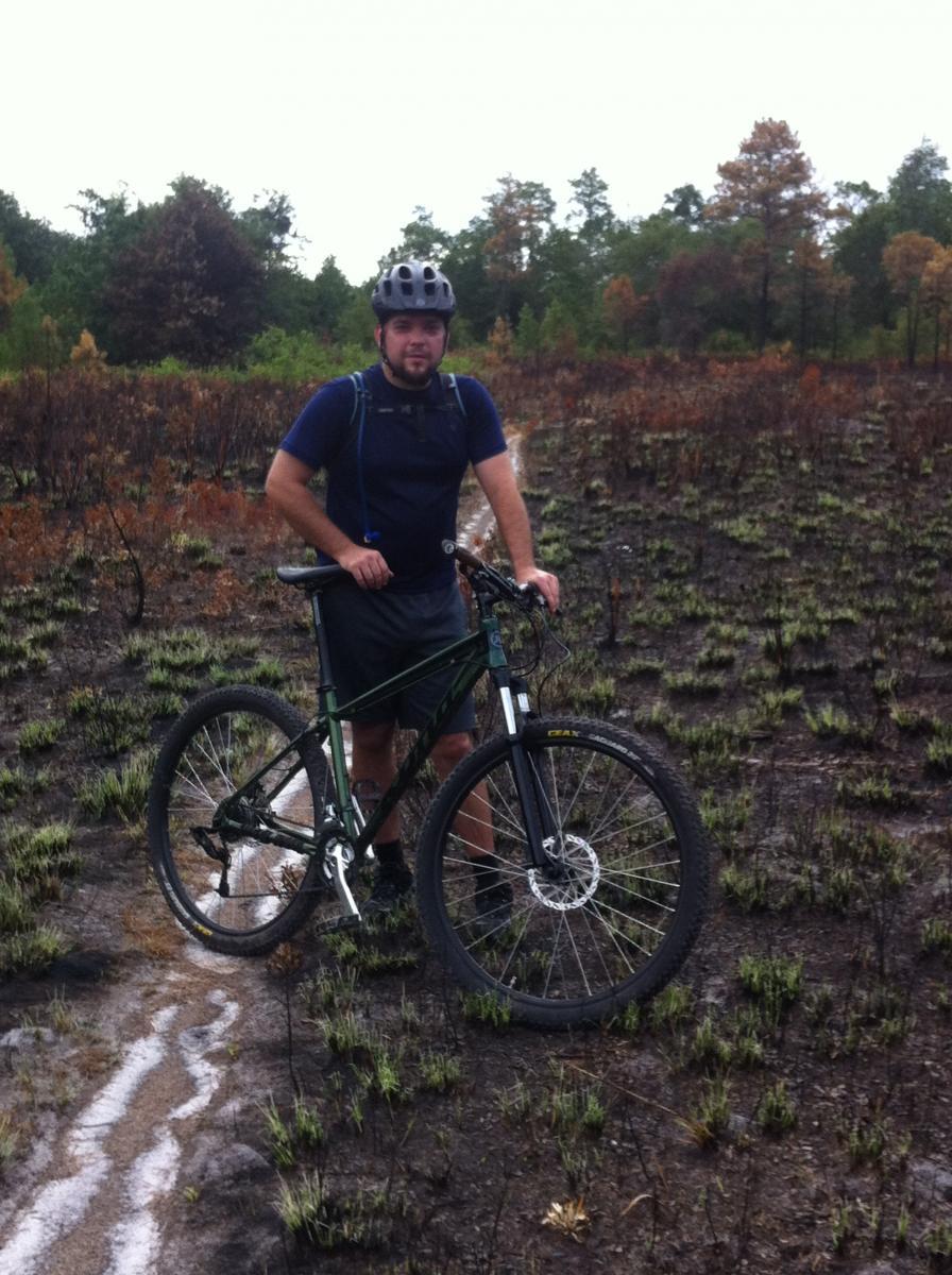 Kona Mahuna: A person in a helmet stands next to a mountain bike on a path through a landscape with burnt vegetation and some regrowth. The sky is overcast, suggesting rainy weather.
