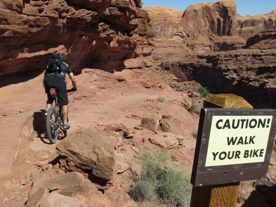 A mountain biker approaches a rocky trail with a caution sign that reads "CAUTION! WALK YOUR BIKE." The landscape features reddish rock formations and sparse vegetation, suggesting a rugged outdoor environment. Captain Ahab mountain bike trail.