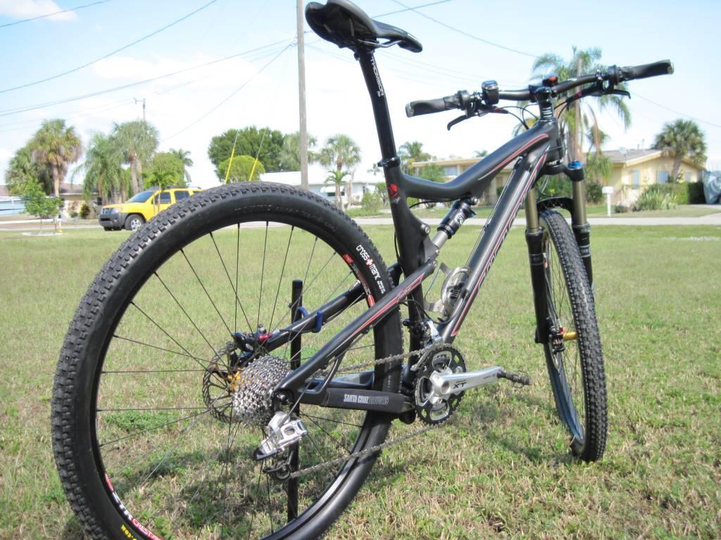 Santa Cruz Tallboy Carbon: A close-up image of a mountain bike parked on a grassy area, showcasing its sleek black frame, geared drivetrain, and thick tires. The bike is positioned at an angle, highlighting its suspension components and handlebars, with a background featuring trees and a yellow vehicle in a residential setting.