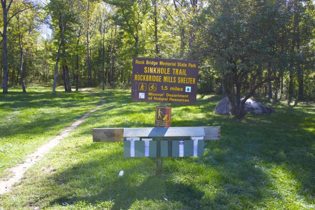 Sign for the Sinkhole Trail at Rock Bridge Memorial State Park, indicating a trail length of 1.5 miles. The sign is surrounded by lush green grass and trees, with a walking path visible leading into the park. Rock Bridge Memorial State Park mountain bike trail.