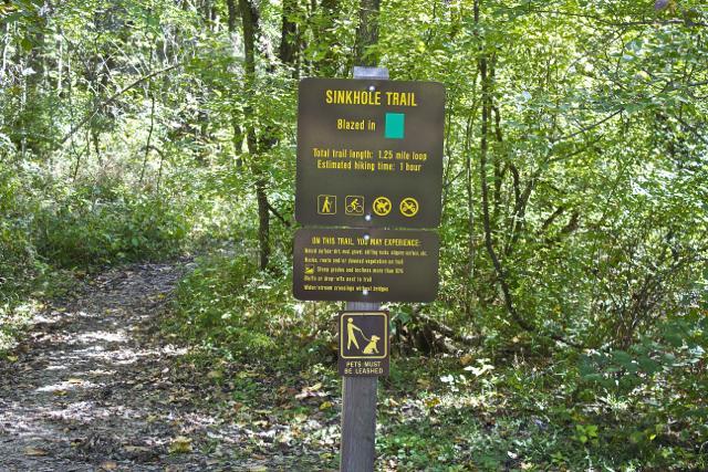 A sign marking the Sinkhole Trail, providing information about the trail length of 1.25 miles, estimated hiking time of 1 hour, and indicating that pets must be leashed. The sign is situated along a wooded path with greenery surrounding it. Rock Bridge Memorial State Park mountain bike trail.