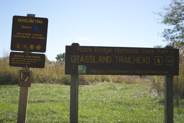 Signage at the Grassland Trailhead in Rock Bridge Memorial State Park. The signs provide information about the trail length (2 miles), estimated hiking time (30 minutes), and include various symbols for trail use and guidelines. The surrounding area features tall grass and trees. Rock Bridge Memorial State Park mountain bike trail.