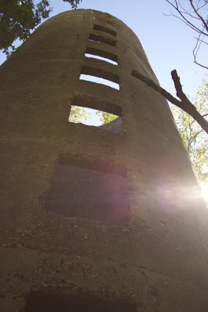 A tall, weathered silo stands against a clear sky, with sunlight illuminating its textured surface. The silo features several rectangular openings along its side, surrounded by trees that frame the scene. Rock Bridge Memorial State Park mountain bike trail.