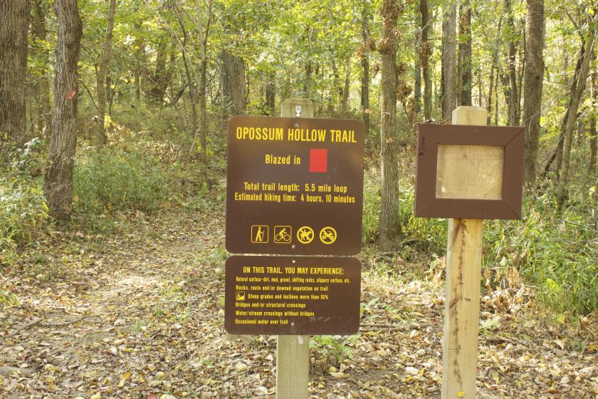 Sign indicating the Opossum Hollow Trail, a hiking path marked with red blazes. The sign details the trail's length (5.5 miles) and estimated hiking time (4 hours, 10 minutes), along with warnings about trail conditions and possible experiences such as rocky terrain, overgrown vegetation, and water crossings. The trailhead is surrounded by trees and natural foliage. Opossum Hollow mountain bike trail.