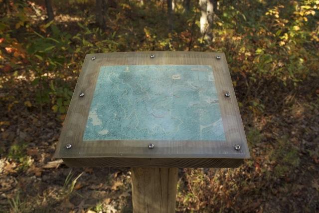 A wooden sign post with a map display on top, surrounded by trees in a forest during autumn. The map is mounted on the post and shows a blue-green landscape, indicating trails and features in the area. The surrounding foliage features orange and yellow leaves, suggesting a seasonal setting. Opossum Hollow mountain bike trail.
