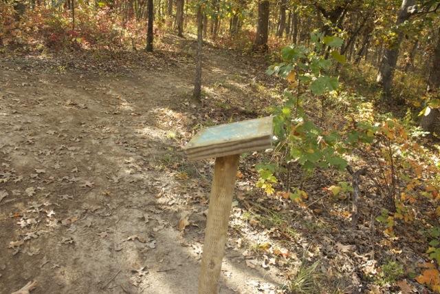 A wooden signpost with a weathered plaque stands on a dirt path surrounded by autumn foliage in a forest. The pathway forks ahead, leading into different areas of the wooded landscape. Opossum Hollow mountain bike trail.