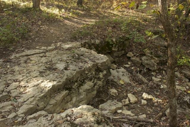 Rocky terrain with a flat, exposed limestone surface surrounded by wooded area and fallen leaves, indicating an autumn setting. A narrow dirt path leads into the background. Opossum Hollow mountain bike trail.
