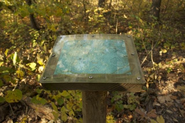 A weathered information sign mounted on a wooden post, displaying a faded map. The sign is surrounded by autumn foliage and forest undergrowth. Opossum Hollow mountain bike trail.