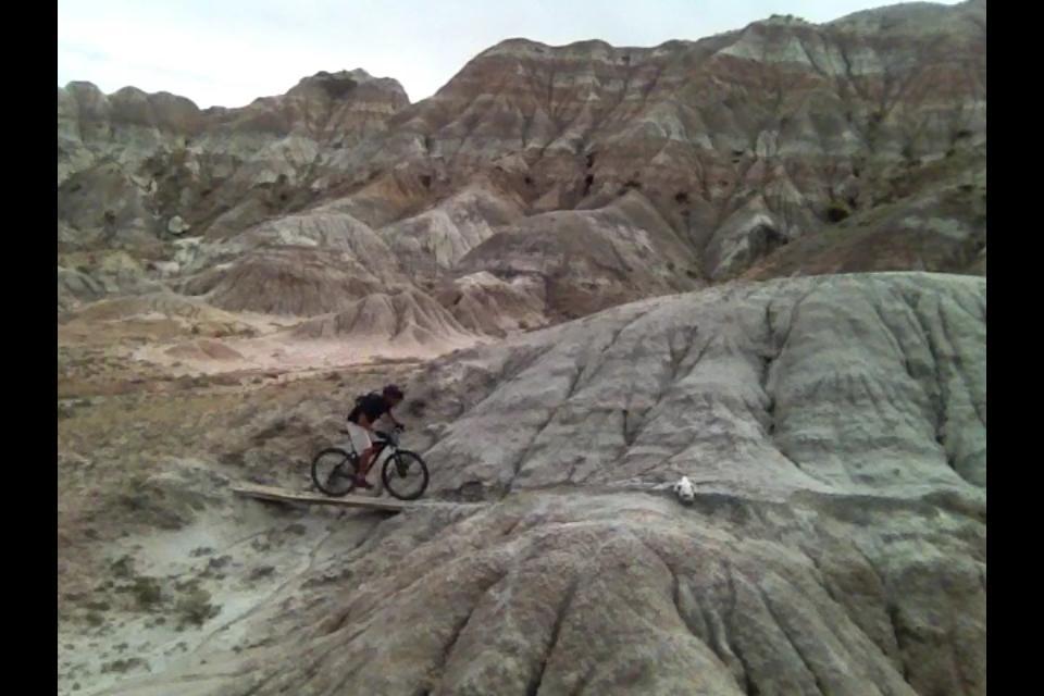 A mountain biker navigating a wooden bridge over rugged, colorful terrain in a desert landscape, with layered rock formations in the background. En Tus Suenos mountain bike trail.