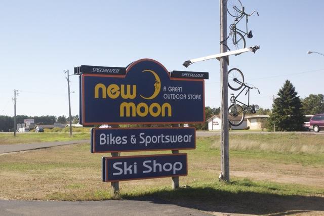 Sign for "New Moon," an outdoor store featuring "Bikes & Sportswear" and a "Ski Shop." The sign displays the store's name prominently, accompanied by a graphic of a moon, and bicycles are mounted above it. The setting is a grassy area beside a road.