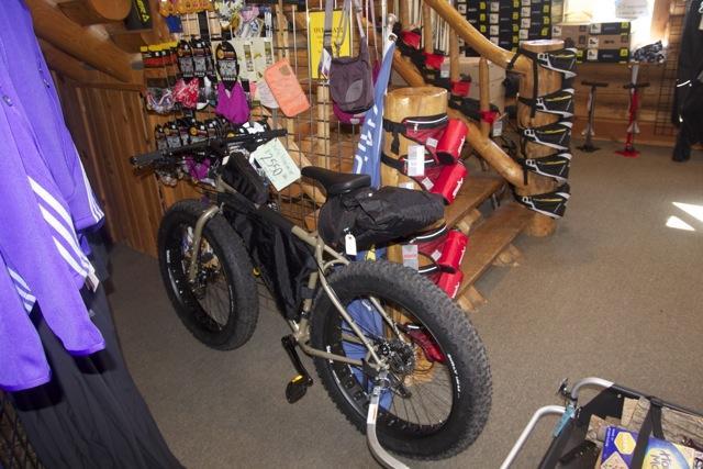 A fat bike displayed in an outdoor sports shop, featuring oversized tires and attached gear. The background includes clothing items and various biking accessories displayed on racks, with wooden accents in the store